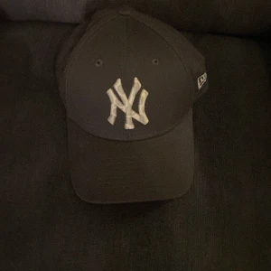 Svart keps från New Era - Snygg svart keps från New Era med det klassiska NY-logotypen framtill. Kepsen har en justerbar rem baktill för perfekt passform och är en del av 9FORTY-serien.