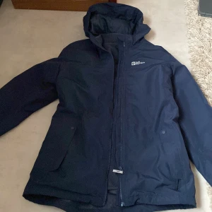 Mörkblå jacka från Jack Wolfskin - Säljer en mörkblå jacka från Jack Wolfskin med huva och dragkedja. Perfekt för kyligare dagar. Jackan har en stilren design med diskret logga på bröstet.