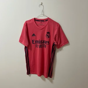 Real Madrid fotbollströja - Snygg rosa fotbollströja från Adidas med Real Madrid-logga och 'Emirates Fly Better' tryckt på framsidan. Tröjan har korta ärmar och mörkblå detaljer längs sidorna. Perfekt för fotbollsfans som vill sticka ut på planen! ⚽