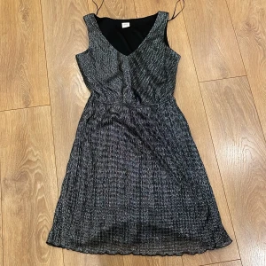 Svart glittrig klänning från Esprit - Snygg svart glittrig klänning från Esprit med v-ringning och ärmlös design. Perfekt för festliga tillfällen med sitt skimrande material.