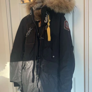 Parajumper Woman - Säljer en svart parkajacka från Parajumpers med pälsfodrad huva och flera praktiska fickor. Jackan har en gul dragkedjedetalj och en patch på ärmen. Perfekt för kalla vinterdagar.