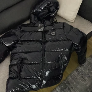 Svart pufferjacka från Trapstar - Säljer en svart, glansig pufferjacka från Trapstar. Jackan har en dragkedja och en huva, perfekt för kyliga dagar. Den finns i alla storlekar och har en modern look som passar bra till streetwear-stilen.