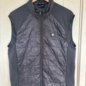 Grå quiltad väst från EA7 Emporio Armani - Säljer en snygg grå quiltad väst från EA7 Emporio Armani. Västen har en dragkedja framtill och ett stilrent märke på bröstet. Perfekt för lager-på-lager under höst och vår. Den är i bra skick och redo för nya äventyr!