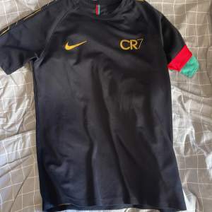 Säljer en svart CR7 t-shirt från Nike i storlek L. Den har korta ärmar och är gjord i Dri-FIT-material, perfekt för träning. T-shirten har guldiga detaljer och en röd och grön rand på ena ärmen. Superbekväm och stilren design!