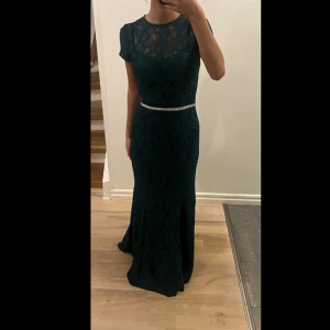 Mörkgrön spetsklänning med bälte - Säljer en elegant mörkgrön långklänning i spets. Klänningen har korta ärmar och en vacker öppen rygg. Den är figursydd med ett glittrigt bälte i midjan som ger en fin silhuett. Perfekt för festliga tillfällen!