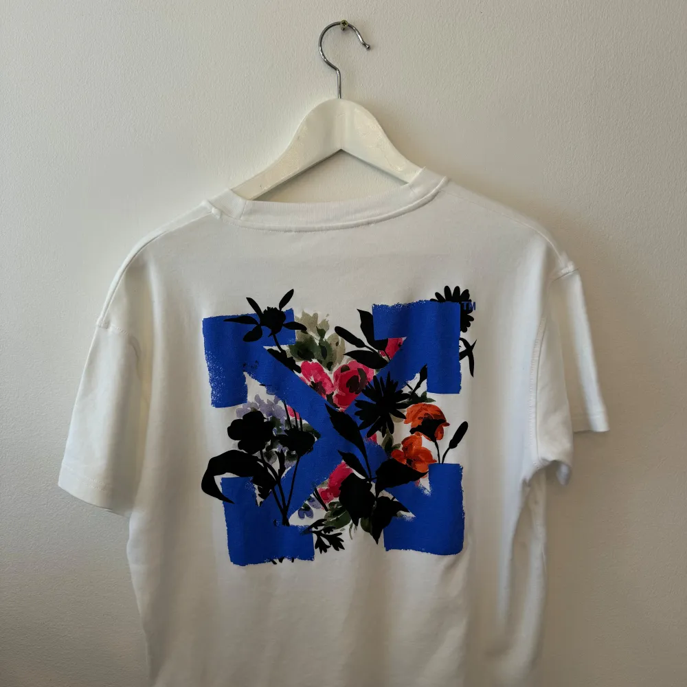 Säljer en snygg vit t-shirt från Off-White i storlek medium. Den har ett coolt blommigt tryck på baksidan med blå detaljer. Perfekt för sommaren och att styla med jeans eller shorts. T-shirten är i bomull och har en normal passform.. T-shirts.