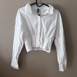 Vit croppad skjorta från FB Sister - Säljer en snygg vit croppad skjorta från FB Sister. Skjortan har långa ärmar och knappar framtill. Den är perfekt för både vardag och fest, med en elastisk midja som ger en fin passform. Materialet känns lätt och bekvämt. Perfekt för vår och sommar! 🌼