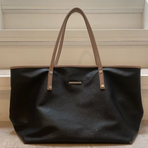 Svart shoppingväska från Fiona Hobo - Säljer en stilren & rymlig svart shoppingväska från Björn Borg. Väskan har bruna handtag och en diskret logga på framsidan. Insidan är fodrad med ett mörkt tyg och har en praktisk innerficka med dragkedja. Perfekt för både vardag och shoppingrundor!