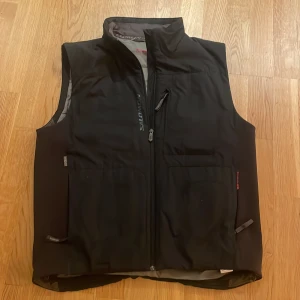 Svart väst från Salomon - Snygg svart väst från Salomon med dragkedja och flera praktiska fickor. Västen är tillverkad med Primaloft-teknologi för extra värme och komfort. Perfekt för kyligare dagar.