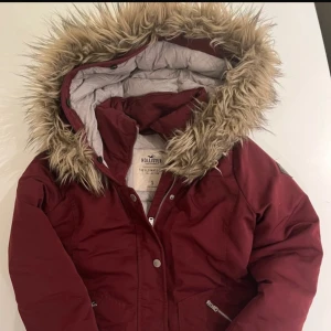 Vinröd dunjacka från Hollister - Säljer en snygg vinröd dunjacka från Hollister med pälskantad huva. Jackan har dragkedja och knappar framtill samt praktiska fickor med dragkedja. Perfekt för kyliga dagar!