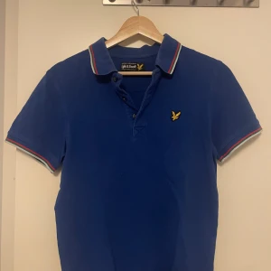 Blå pikétröja från Lyle & Scott - Säljer en snygg blå pikétröja från Lyle & Scott med korta ärmar och klassisk krage. Tröjan har en broderad logotyp på bröstet och ränder i rött och vitt på krage och ärmslut. Perfekt för en stilren look!