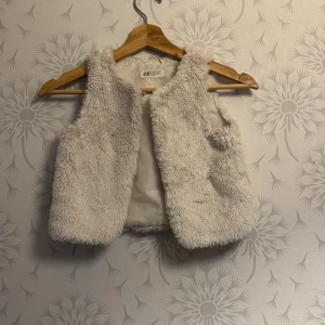 Vit pälsväst från H&M - Säljer en fluffig och mysig vit pälsväst från H&M. Perfekt för att hålla sig varm och stilren under kyligare dagar. Västen är ärmlös och har en mjuk insida.