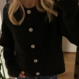 Svart kofta med knappar - Snygg svart kofta med stora silverfärgade knappar framtill. Den har en klassisk design med långärmade ärmar och en normal passform. Perfekt för att ge en stilren look till din outfit.