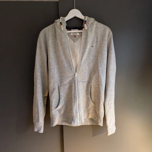 Grå hoodie från Tommy Hilfiger Denim - Säljer en stilren grå hoodie från Hilfiger Denim. Tröjan har en dragkedja framtill och två praktiska fickor. Den är långärmad och har en klassisk passform. Perfekt för en avslappnad look.