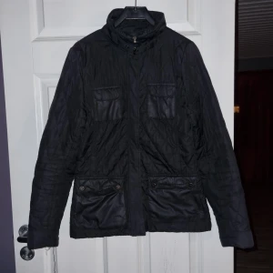 Svart quiltad jacka från H&M - Säljer en svart quiltad jacka från H&M med dragkedja och flera fickor framtill. Jackan har långa ärmar och en stilren design. Perfekt för dig som vill ha en praktisk och snygg jacka i garderoben. Sparsamt använd,  har dock tappat bort midjeskärpet.