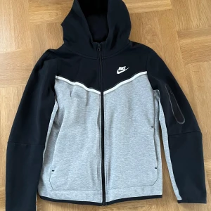 Grå och svart nike tech fleece  - Säljer en snygg grå och svart hoodie från Nike med dragkedja och huva. Hoodien är i storlek xl barn (158-170 cm)