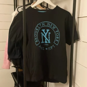 Svart t-shirt med Brooklyn-tryck - Snygg svart t-shirt från WA med ett stort tryck i blått som säger 'Brooklyn New York 1996'. Perfekt för en avslappnad stil. T-shirten har en normal passform och korta ärmar.