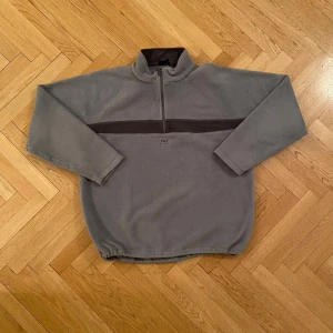 Helly Hansen Fleece Tröja - Säljer en grå/grön fleece tröja från Helly Hansen med halv dragkedja och mörkare grått band över bröstet. Tröjan har långa ärmar och en hög krage samt justerbar öppning i midjan. Det står XXL men den passar ungeför som XL. Perfekt för skön oversized fleece