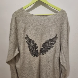 Grå tröja med vingmotiv från Hunkydory - Säljer en stilren grå tröja från Hunkydory med ett coolt vingmotiv på ryggen. Tröjan har en rund hals och långa ärmar, perfekt för en avslappnad look. Passar bra till både jeans och kjol. Angora, lammull och lite polyester.