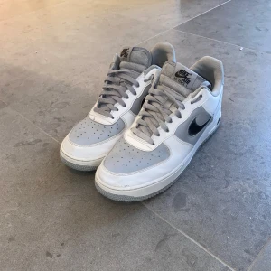 Nike Air Force 1 White Wolf Grey Black - Snygga Nike Air Force sneakers i vitt och grått med klassisk design. Skorna har en svart Nike-logga på sidan och grå snörning. Perfekta för en stilren look.