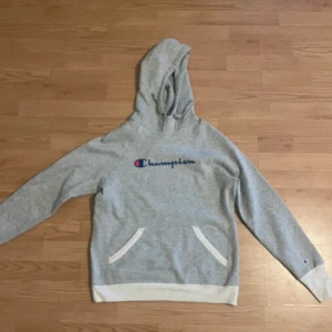 Grå hoodie från Champion - Säljer en grå hoodie från Champion med deras klassiska logga broderad på bröstet. Tröjan har en känguruficka och ribbade muddar vid ärmslut och nederkant. Perfekt för en avslappnad stil.