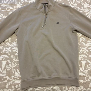 Beige tröja från Calvin Klein Jeans - Säljer en stilren beige tröja från Calvin Klein Jeans med dragkedja vid halsen och broderad logga på bröstet. Tröjan har långa ärmar och ribbade muddar för en bekväm passform. Perfekt för en avslappnad look.