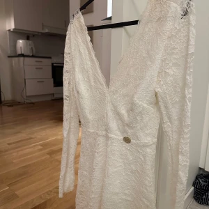 Vit spetsklänning från Chiara Forthi - Elegant vit spetsklänning från Chiara Forthi med v-ringad design och långa ärmar. Klänningen har en fin spetsdetalj och en liten metallbricka med märkeslogga i midjan. Perfekt för speciella tillfällen.