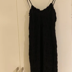 Svart klänning från H&M - Snygg svart klänning från H&M med smala axelband och en rynkad textur. Perfekt för en stilren look. Klänningen har en enkel design som gör den lätt att matcha med olika accessoarer. Tjusterbara band