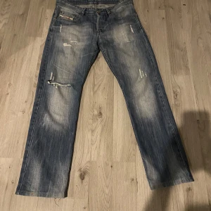 Blå jeans från Diesel - Säljer ett par blå jeans från Diesel med slitna detaljer och raka ben. Jeansen har en klassisk femficksdesign och en läderpatch med Diesel-logga på baksidan. Perfekta för en avslappnad stil.