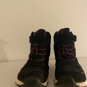 Svarta boots från Treksta - Svarta boots från Treksta med lila detaljer och snörning. De har en hög modell med robust sula, perfekt för utomhusaktiviteter. Märket Treksta är synligt på sidan av skorna.