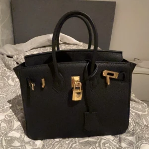 Svart handväska med gulddetaljer - Elegant svart handväska med gulddetaljer och lås. Perfekt för att ge en lyxig touch till din outfit. Väskan har en klassisk design med två handtag och en strukturerad form.