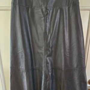 Svart långkjol i skinnimitation från Lindex - Snygg svart långkjol i skinnimitation från Lindex. Kjolen har en stilren design med sömmar som ger en elegant touch. Perfekt för att skapa en chic look.