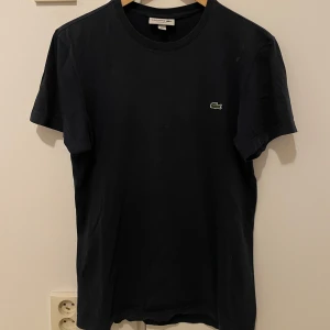 Svart t-shirt från Lacoste - Snygg svart t-shirt från Lacoste med det klassiska gröna krokodilmärket på bröstet. T-shirten har en normal passform och är tillverkad i mjukt bomullsmaterial. Perfekt för en stilren och avslappnad look.