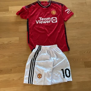 Röd Manchester United fotbollströja med shorts - Säljer en röd Manchester United fotbollströja med svarta detaljer och TeamViewer-logga. Tröjan har korta ärmar och ett klubbmärke på bröstet. Tillhörande vita shorts med svarta ränder och nummer 10. Perfekt för fans! ⚽