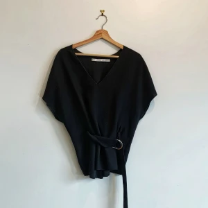 Svart topp från & Other Stories - Snygg svart topp från & Other Stories i storlek S. Den har en v-ringad design och ett bälte i midjan för en stilren look. Perfekt för att ge en elegant touch till din outfit.