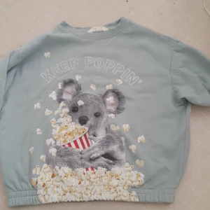 Ljusgröntröja med koalamotiv från H&M - Säljer en ljusblå tröja från H&M med ett gulligt koalamotiv som håller en popcornhink och texten 'Keep Poppin'. Tröjan har långa ärmar och en rund halsringning. Perfekt för en avslappnad stil!