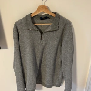 Grå tröja med dragkedja från Polo Ralph Lauren - Säljer en stilren grå tröja från Polo Ralph Lauren. Tröjan har en halv dragkedja och långa ärmar, perfekt för en avslappnad look. Den är tillverkad i ett mjukt material som ger en bekväm passform. Storleken är M.