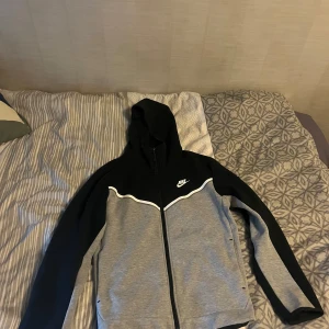 Svart och grå huvtröja från Nike - Säljer en snygg Nike tech fleece från Nike i svart och grått. Knappt använd och i bra skick. Tröjan har en dragkedja framtill och en klassisk Nike-logga på bröstet. Perfekt för en sportig look eller avslappnad stil. 