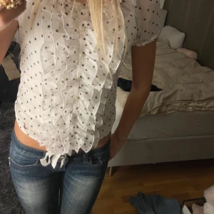 Vit prickig blus med volangdetaljer - Charmig vit blus med svarta prickar och volangdetaljer framtill. Blusen har korta ärmar och en söt knytning vid midjan. Perfekt för en stilfull och lekfull look.