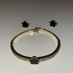 Guldarmband och örhängen med svart blomma - Elegant set med ett guldarmband och matchande örhängen. Armbandet har en svart blomma i mitten och är dekorerat med gnistrande stenar längs sidorna. Örhängena har samma svarta blomdesign, vilket ger en stilren och sammanhängande look.