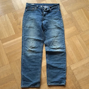 Blå jeans från Levi's - Säljer ett par klassiska blå jeans från Levi's med en loose passform. De har en traditionell femficksdesign och en snygg slitning för en avslappnad look. Perfekta för en casual stil.