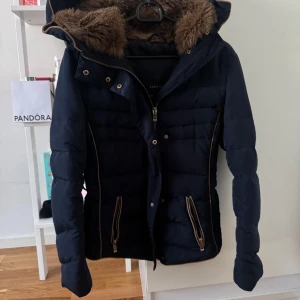Mörkblå dunjacka med pälskrage - Snygg mörkblå dunjacka från Zara med avtagbar huva och pälskrage. Jackan har dragkedja och knappar framtill samt praktiska fickor med dragkedja. Perfekt för kyliga dagar.