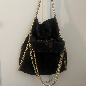 Stella mcartney liknande  - Snygg svart axelväska med glittrig yta och guldiga kedjor som axelremmar. Väskan har en unik design med en stor öppning och dekorativa kedjor som ger en lyxig känsla. Perfekt för att lyfta vilken outfit som helst.