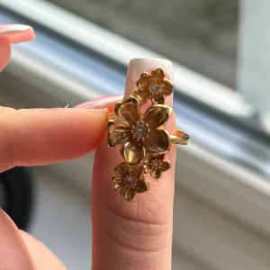 Guldfärgad blomring - Elegant guldfärgad ring med blommotiv och små gnistrande stenar i mitten av varje blomma. Perfekt för att ge en touch av glamour till din outfit. (Den är i rostfritt stål ifrån Lyla, men jag har lackat över den med nagel lack som är genomskinlig) Använd en gång