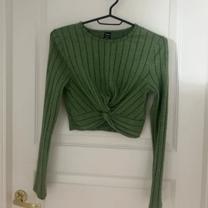 Grön ribbad tröja från H&M - Snygg grön ribbad tröja  med en twistdetalj framtill. Tröjan är croppad och har långa ärmar, helt oanvänd! 