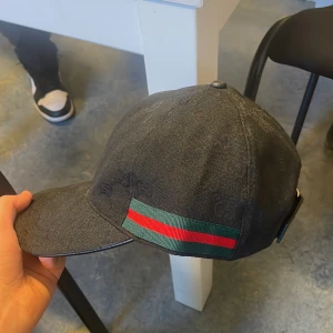 Svart keps från Gucci - Snygg svart keps från Gucci med diskret mönster och klassiska gröna och röda ränder på sidan. Justerbar rem baktill för perfekt passform. Perfekt för en stilren look.