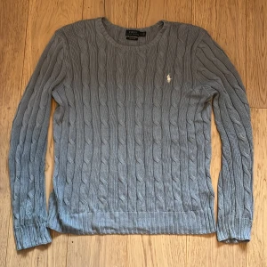 Grå stickad tröja från Polo Ralph Lauren - Snygg grå stickad tröja från Polo Ralph Lauren med kabelstickat mönster. Tröjan har långa ärmar och en liten broderad logga på bröstet. Perfekt för kyligare dagar. Köptes för 2 395 säljs för 1 500kr, vet inte direkt storleken, står LG tror det är storlek L