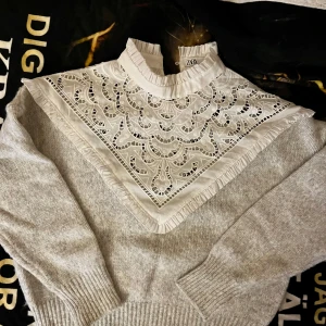 Grå tröja med spetsdetaljer från Zara - Säljer en stilren grå tröja från Zara med vackra spetsdetaljer över bröstet. Tröjan har långa ärmar och ribbade muddar vid ärmslut och nederkant. Perfekt för en elegant look.