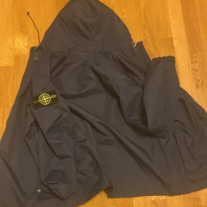 Mörkblå jacka från Stone Island - Säljer en snygg mörkblå jacka från Stone Island med huva och dragkedja. Jackan har en ikonisk logga på ärmen och är perfekt för en stilren look. Den är tillverkad i ett slitstarkt material och har en modern design.