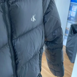 Svart dunjacka från Calvin Klein - Snygg svart dunjacka från Calvin Klein med dragkedja och huva. Jackan har långa ärmar och två fickor framtill med knappar. Perfekt för kyliga dagar.